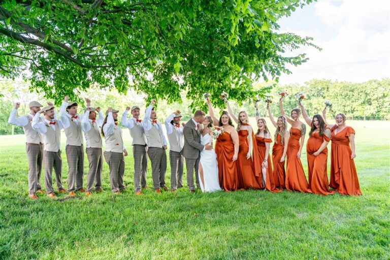 Full wedding party in rust dresses and tan vests raising bouquets and fists under a large tree at Tobacco Barn Farm in Holt, Missouri