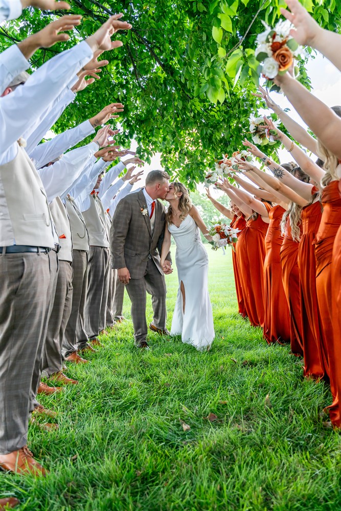 Newlyweds kissing at the end of a tunnel of raised arms formed by their wedding party under a large shade tree at Tobacco Barn Farm