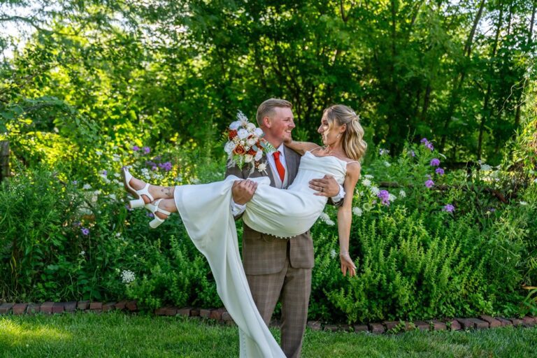 Groom in a brown plaid suit carrying his bride in his arms in a lush green garden at Tobacco Barn Farm in Holt, Missouri
