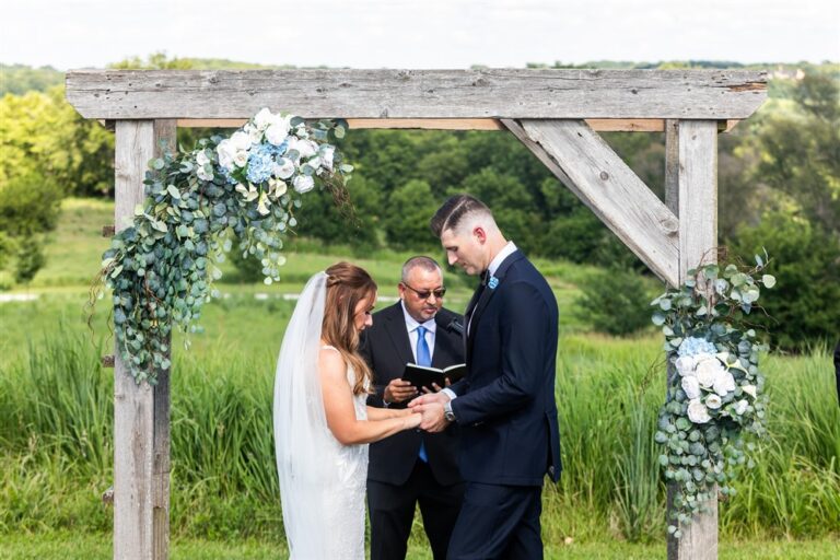 Bride and groom exchange rings under a floral wooden arbor during their outdoor ceremony at Red Barn Farm in Weston, Missouri