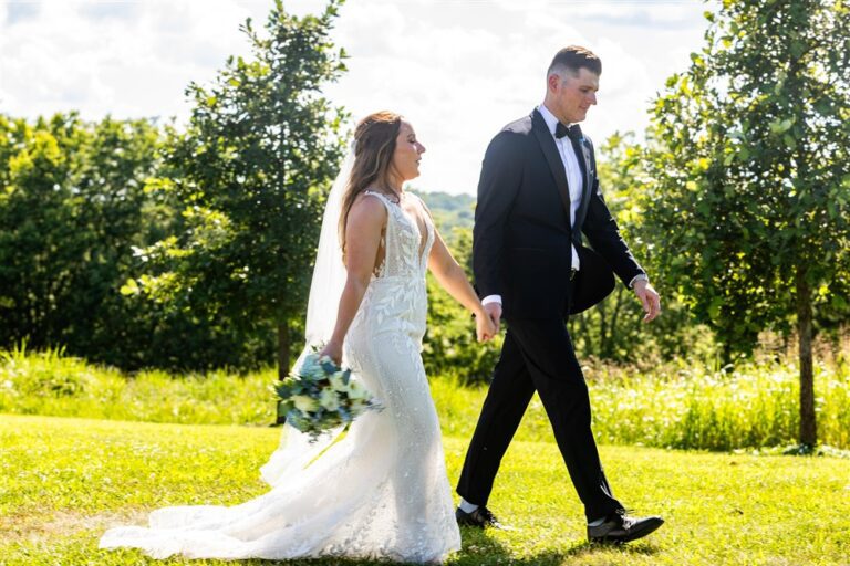 Bride and groom walk hand in hand across a sunlit orchard lawn at Red Barn Farm wedding venue in Weston, Missouri