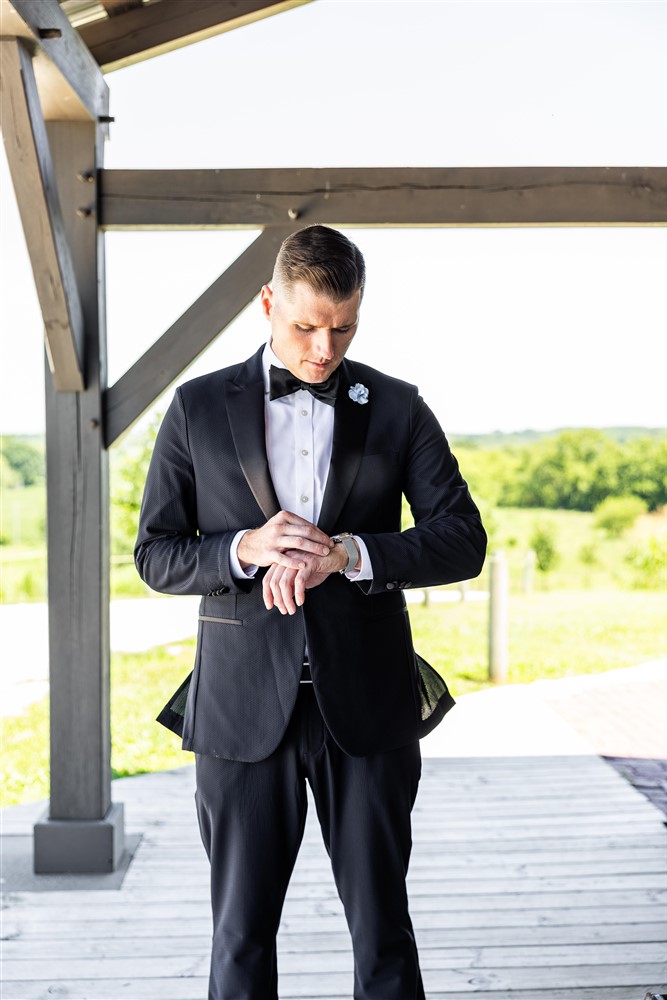Groom in a black tuxedo adjusts his watch before the ceremony at Red Barn Farm in Weston, Missouri