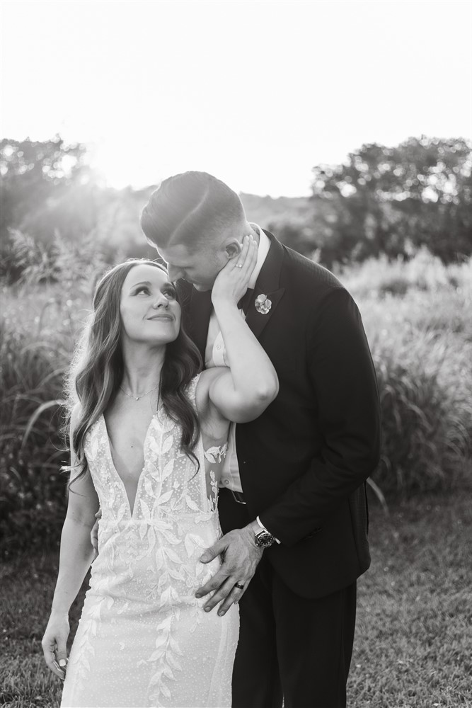 Black and white portrait of a bride touching the groom's face at sunset in a field at Red Barn Farm in Weston, Missouri