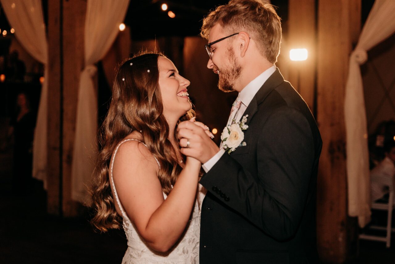 Bride and groom smile and hold hands during their first dance at a warmly lit indoor wedding reception, captured by Complete Weddings + Events