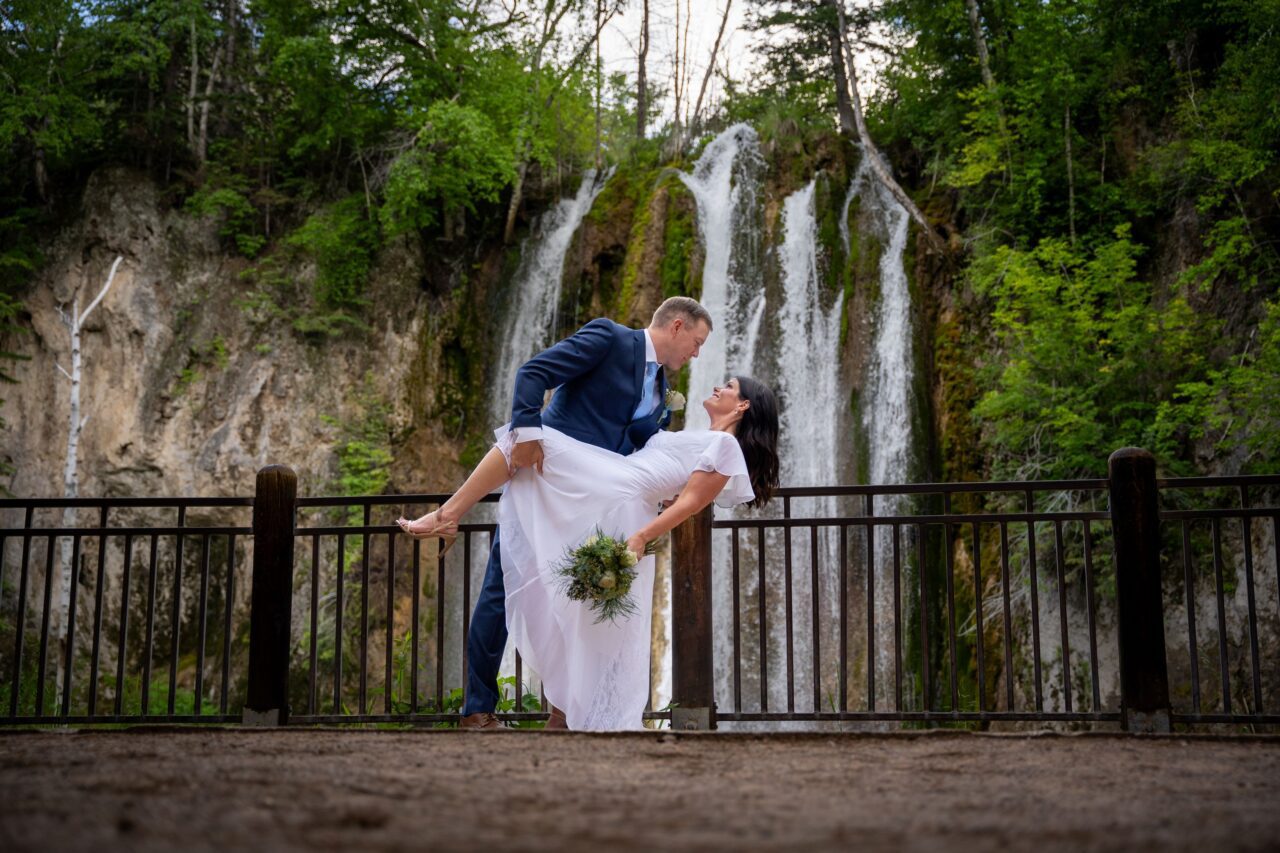 A groom in a dark blue tuxedo dips a bride who is wearing a long, flowing wedding dress and holding her bouquet down to her side. Behind them is a natural waterfall.