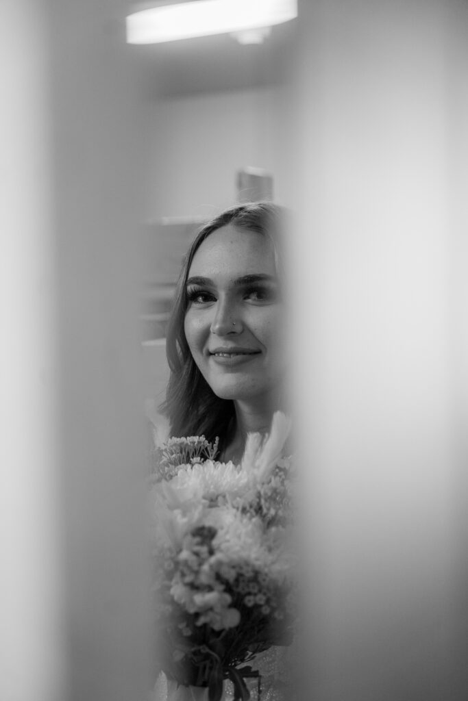 A candid photo of a bride, taken between two doors while she holds her bouquet and smiles.