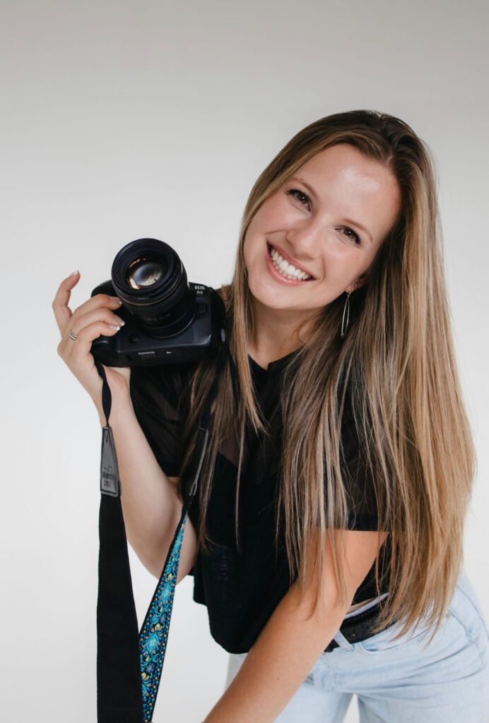 This image shows Anna Mahoney, the lead wedding content creator at Complete Weddings and Events. She has long blonde hair and is holding her DSLR camera while smiling.