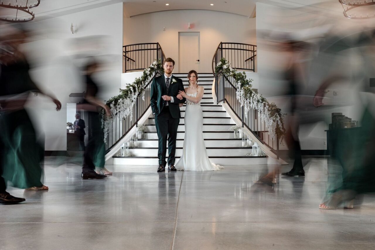 Portrait of a bride and groom at the bottom of a grand staircase. Wedding party members pass by on either side with a motion-blur effect.