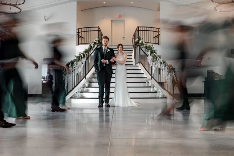 Portrait of a bride and groom at the bottom of a grand staircase. Wedding party members pass by on either side with a motion-blur effect.
