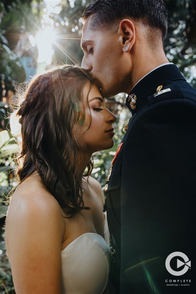 Groom kisses bride on forehead while sun shines through background