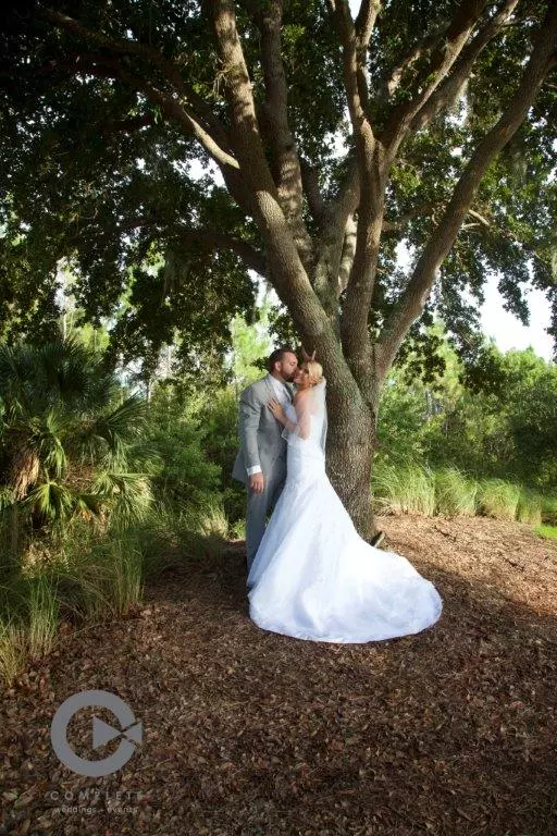 Bride in Wedding Dress and Groom in Suit Embrace under Tree in a photo with a watermark by Complete Weddings + Events