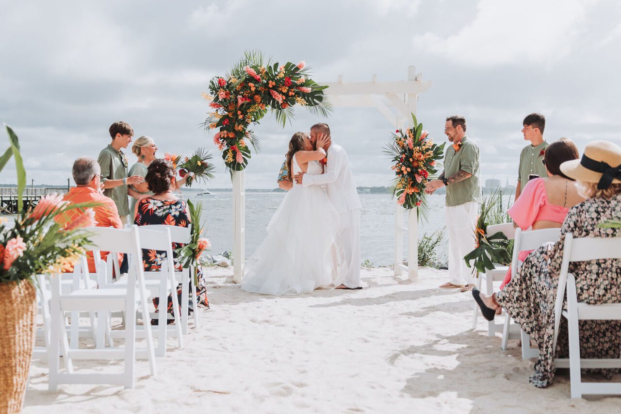 Couple kissing at a beach wedding ceremony framed by floral arches
