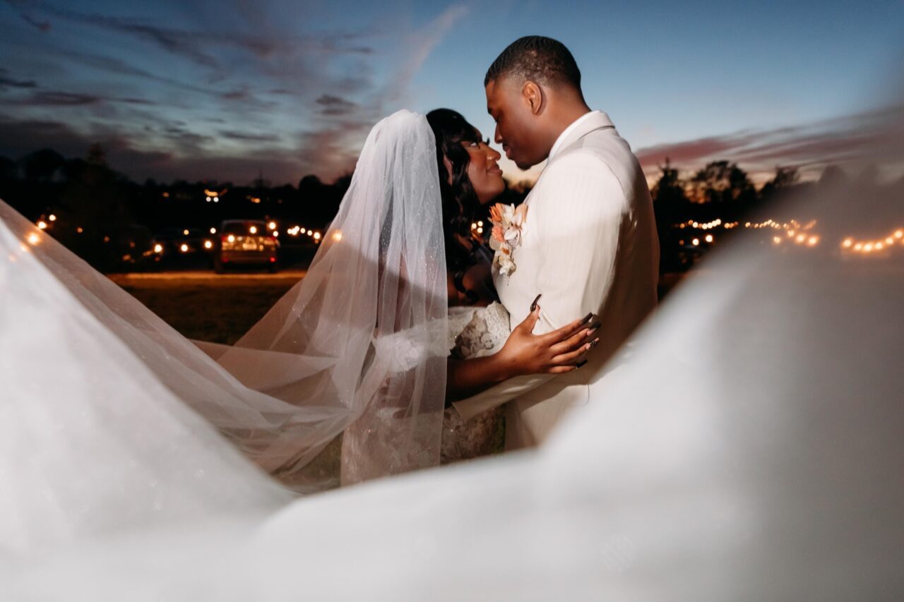 Couple embracing beneath a veil at dusk with warm string lights behind them
