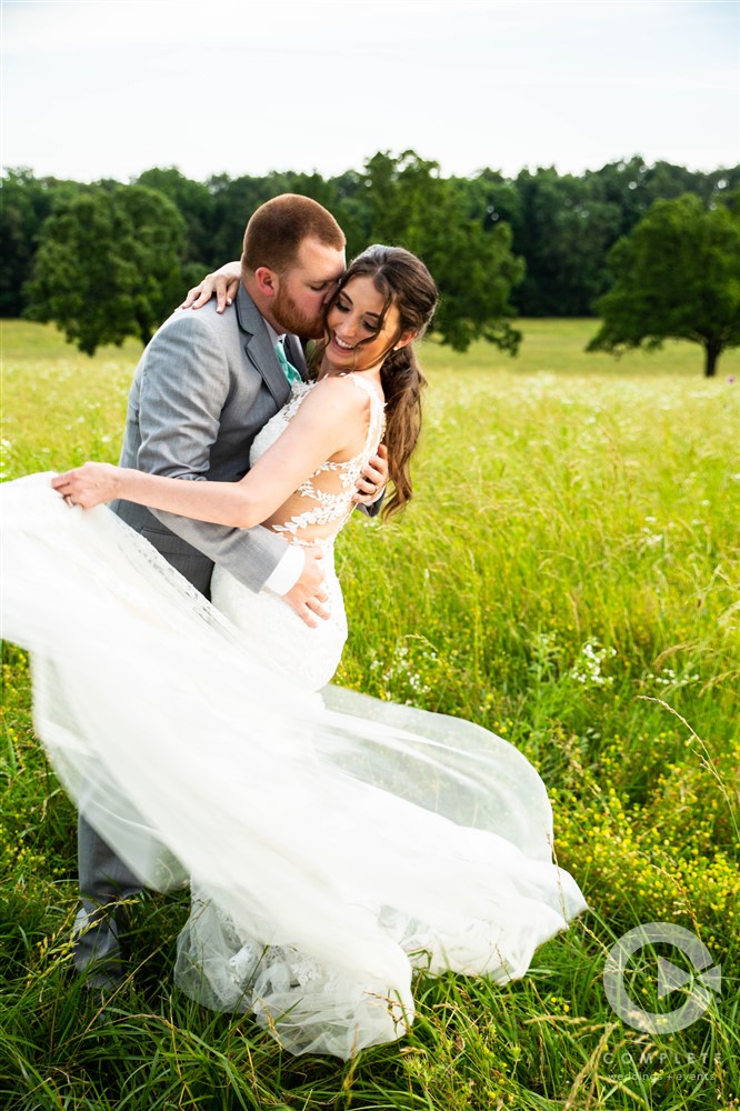 Bride and groom kiss in field while bride plays with dress