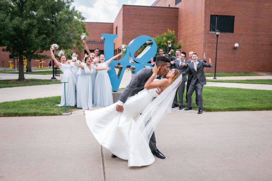 Groom dipping his Bride in front of wedding party and Love sign at the Wichita State University campus, Wichita, KS.